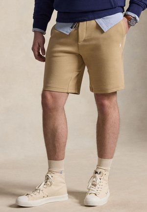 Homme portant un short beige, des baskets montantes beige clair, des chaussettes beiges, un sweat bleu, une chemise bleu clair et une montre-bracelet, debout avec une main dans la poche.