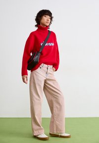 Rode gebreide coltrui met 'essesse' graphic, gecombineerd met lichtroze wijde jeans en een zwarte schoudertas. Witte sneakers met een bruine zool.