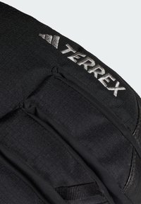 Sort Terrex rygsæk sektion med struktureret stof, buet form og reflekterende sølvbrand. Inkluderer elastiske remme og syede detaljer.