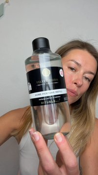 Donna con capelli castano chiaro tiene una bottiglia trasparente etichettata "Velvet Oudh Home Perfume Refill" di Rituals contro una parete semplice.