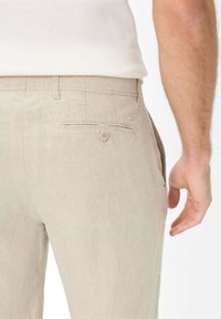 Beige linen trousers featuring a back pocket with a button closure and subtle texture. Side view shows a straight fit and tailored design.