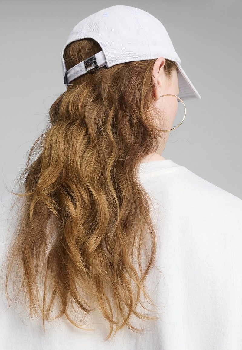 Casquette de baseball blanche avec une visière incurvée, une sangle réglable et un tissu texturé, associée à de longs cheveux bruns ondulés et une chemise blanche.