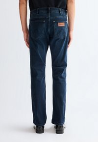 Moški stoji, oblečen v temno modre Wrangler kavbojke ravnega kroja in črne čevlje, viden od zadaj na enobarvni svetli podlagi.