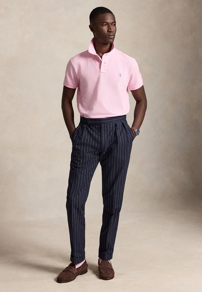 Uomo che indossa una polo rosa chiaro a maniche corte infilata in pantaloni blu navy a righe sottili, con mocassini marroni e un orologio da polso.