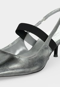 Chaussure argentée à talon kitten avec bout pointu et bride élastique noire sur le cou-de-pied, sur fond uni.