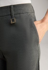 Graue, maßgeschneiderte Hose mit dezenter Textur, einem quadratischen goldenen Akzent am Bund und seitlichen Taschen.