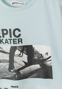 Jasnoniebieska koszula z czarnym napisem "EPIC SKATER" oraz czarno-białym zdjęciem skateboardzistów w akcji w skateparku.