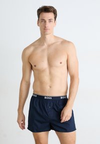 Meztelen férfi rövid barna hajjal és kék szemekkel, sötétkék BOSS boxer alsót viselve, egy sima fehér háttér előtt áll.