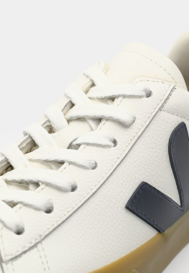 Baskets en cuir blanc avec lacets plats blancs, accent de logo bleu marine et surface texturée. Présente une semelle en caoutchouc et des détails de couture contrastés.