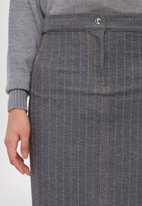 Jupe à fines rayures grises avec surpiqûres brunes et bouton argenté, portée avec un pull gris côtelé à manches longues ; main gauche détendue le long du corps.