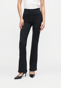 Pantaloni evazați negri din material elastic, cu talie înaltă. Au o textură netedă și un design mulat, asortați cu pantofi negri cu toc înalt.