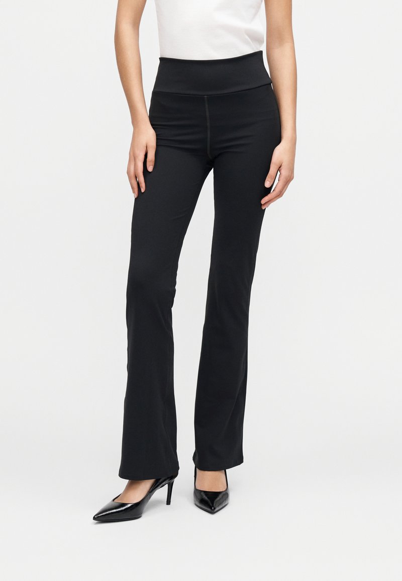 Pantaloni evazați negri din material elastic, cu talie înaltă. Au o textură netedă și un design mulat, asortați cu pantofi negri cu toc înalt.