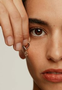 Pendiente de aro en tono dorado con un charm en forma de 'S' adornado con strass, sostenido por una mano cerca de la cara sobre un fondo neutro.