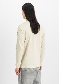 Camisa beige de manga larga hecha de algodón, con cuello redondo y gráficos impresos a lo largo de la manga izquierda, combinada con pantalones gris claro.