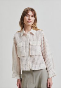 Camisa ligera de color crema con sutiles rayas verticales, que presenta dos bolsillos en el pecho y un cuello clásico, mangas largas con puños abotonados.