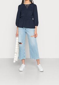 Blusa navy con maniche a sbuffo e vita aderente, abbinata a jeans a gamba larga chiari e distressed e sneakers platform bianche. Borsa bianca con borchie.