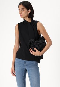 Blusa preta sem mangas com detalhe amarrado no pescoço, combinada com jeans azuis. Segurando uma bolsa de palha preta texturizada.