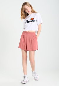 Weißes, kurzes T-Shirt mit schwarzem "Ellesse"-Logo, kombiniert mit rosa Falten-Shorts. Das Model trägt weiße Sneakers und steht vor einem hellen Hintergrund.