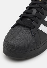 Černá kožená teniska s texturovaným povrchem, výraznou špičkou, černými tkaničkami a bílými pruhy. Jemné zlaté detaily na jazyku.