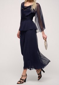 Mörkblå, fladdrande klänning med genomskinliga långärmar och spetsdetaljer på bodicen. I kombination med svarta strap-on klackar och en glittrande clutch.