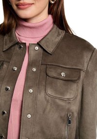 Veste en suède marron avec des boutons-pression, deux poches à l'avant et une poche latérale zippée, superposée à un pull à col roulé rose.