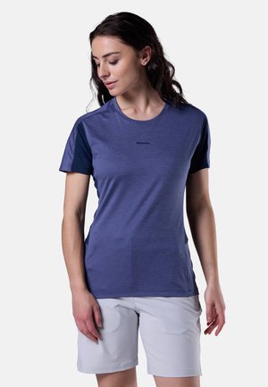 Femme portant un t-shirt violet ajusté à manches courtes et un short gris clair arrivant aux genoux, regardant vers sa gauche en baissant les yeux.