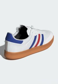 Sneaker bianco con strisce blu e rosse, materiale texturizzato, punta arrotondata e suola in gomma. Il tallone presenta il testo "VELOSAMBA".