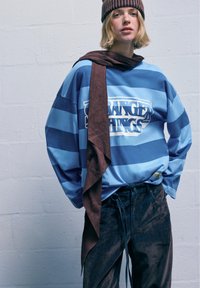 Modrá pruhovaná košeľa s dlhým rukávom a grafikou "STRANGER THINGS", skombinovaná s tmavými, textúrovanými nohavicami a hnedým šálom. Nosí sa s hnedou čiapkou.