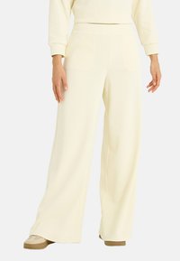 Pantalones de pierna ancha hechos de tela suave y texturizada en un color crema claro. Presentan una cintura alta y un ajuste relajado sin cierres visibles.