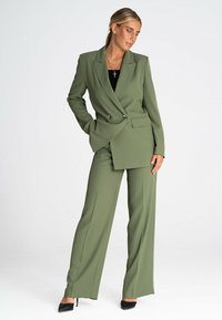 Oliwkowy garnitur skrojony na miarę z podwójnym żakietem i szerokimi spodniami. Zawiera wyraźne klapy i boczne kieszenie. Czarna bluzka i szpilki.