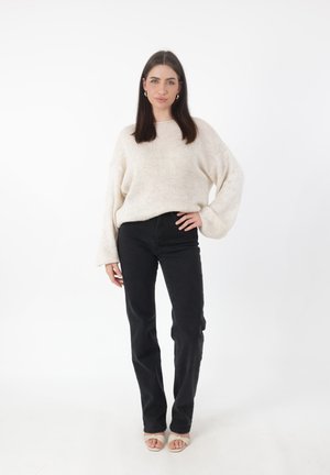 Suéter de punto beige claro con mangas anchas, combinado con jeans negros de pierna recta y sandalias de tacón abierto en color marrón. Fondo blanco liso.