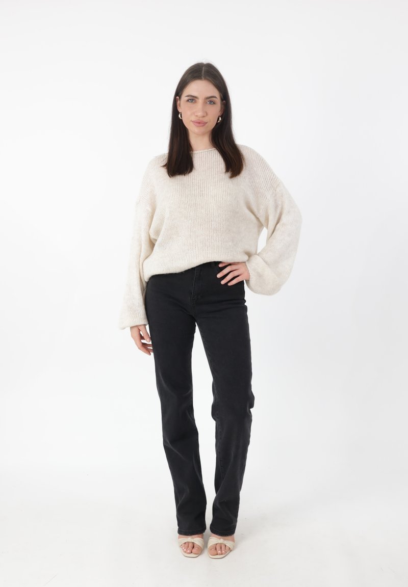 Maglione lavorato a maglia color beige chiaro con maniche ampie, abbinato a jeans neri a gamba dritta e sandali aperti con tacco color sabbia. Sfondo bianco semplice.