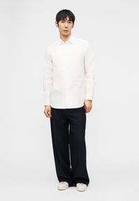 Άνδρας που στέκεται με ουδέτερη έκφραση φορώντας λευκό πουκάμισο με κουμπιά, φαρδιά μαύρα παντελόνια και λευκά παπούτσια μπροστά σε απλό φόντο.