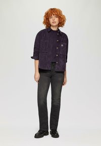 Paarse corduroy shirt jacket met twee voorzakken, zwarte trui eronder, gecombineerd met zwarte jeans en stevige zwarte laarzen.