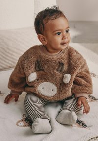 Småbarn som sitter på en djurmönstrad filt, klädd i en brun, lurvig tröja med ett renansikte och randiga leggings.