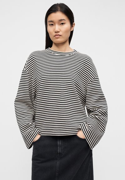 Μπλούζα πλεκτή ριγέ σε μαύρο και κρεμ, με στρογγυλή λαιμόκοψη, μακριά μανίκια και άνετη εφαρμογή, φορεμένη με σκουρόχρωμη τζιν φούστα.