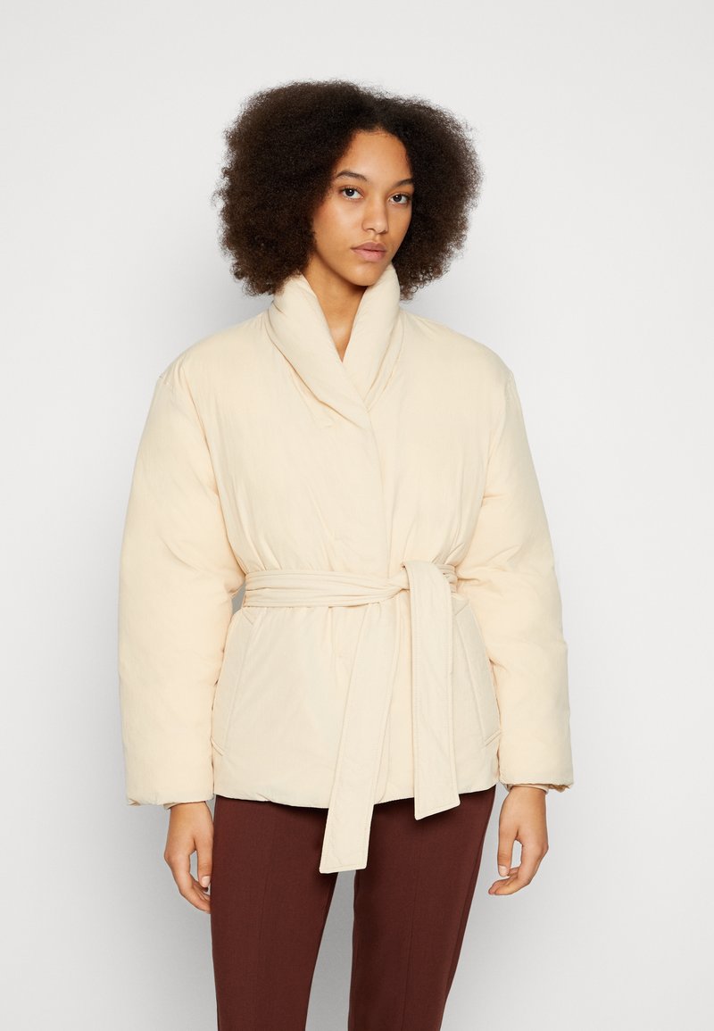 Mulher com cabelo encaracolado vestindo uma jaqueta puffer creme com cinto e calças castanhas, em um fundo branco simples.