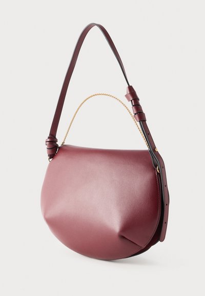 Στρογγυλή τσάντα ώμου από δέρμα μπορντό με ρυθμιζόμενο ιμάντα ώμου και μικρή λαβή με χρυσή αλυσίδα, εμφανίζεται πάνω σε λευκό φόντο.