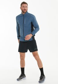 Letvægts blå jakke med sorte detaljer, frontlynlås og reflekterende elementer; parret med sorte shorts og grå sneakers.