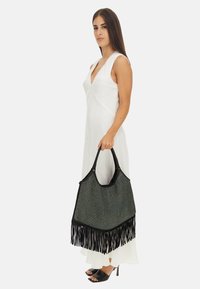 Grande borsa intrecciata nera e verde con fondo frangiato, dotata di manico liscio e dettagli in hardware dorato. Modella in piedi con abito e tacchi.