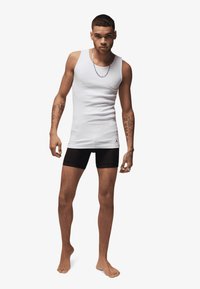 White tank top made of cotton, paired with black fitted shorts. Notable tattoos on arms. Barefoot stance against a plain background.