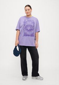 Camiseta oversized morada con gráfico de sol, de mangas cortas, combinada con jeans anchos negros y un bolso texturizado azul marino. Zapatillas plateadas mostradas.