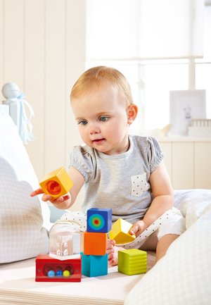 Colorful wooden building blocks in various shapes stacked on a surface; a child holds a yellow square block. Soft backgrounds with light filtering in.