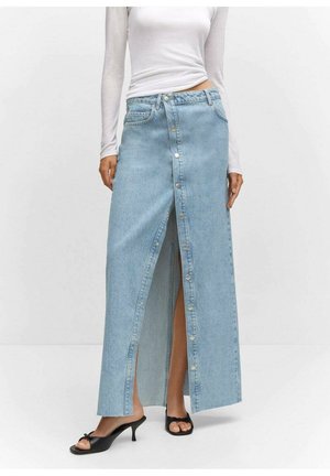 Donna che indossa una lunga gonna di jeans azzurro chiaro con bottoni frontali, top bianco a maniche lunghe e sandali neri con tacco e punta aperta, in piedi su sfondo bianco.