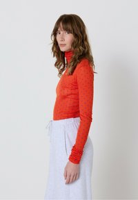 Sieviete ar viļņainiem brūniem matiem, tērpta oranžā punktiņotā augšpusē un gaišajos pelēkos biksēs ar aukliņu, stāv sānis pret vienkāršu fona.