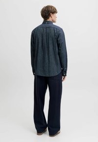 Vīrietis tērpies tumši zila garām piedurknēm krekla un brīvām melnām biksēm, stāv pretēji pret vienkāršu baltu fonu.