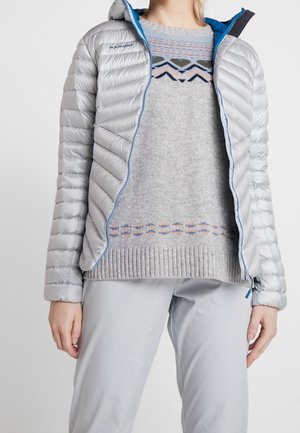 Persoon draagt een lichtgrijs gewatteerd jack met blauwe ritstrim over een gebreide trui met patroon in grijstinten en lichtgrijze broek.