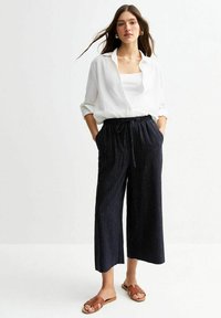 Chemise blanche à boutons et pantalon large à rayures sombres avec poches. Sandales brunes à enfiler. Tissu lisse et coupe décontractée.