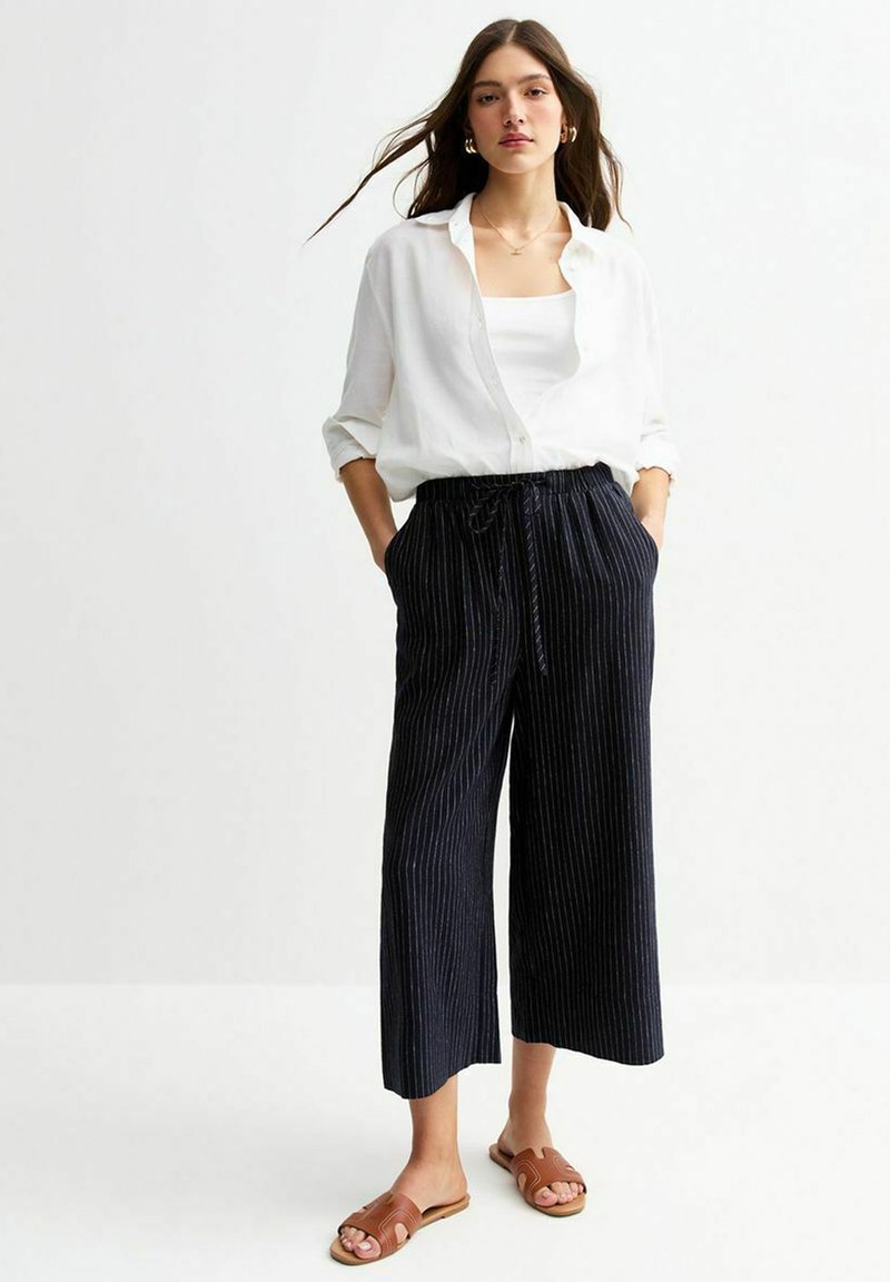 Chemise blanche à boutons et pantalon large à rayures sombres avec poches. Sandales brunes à enfiler. Tissu lisse et coupe décontractée.
