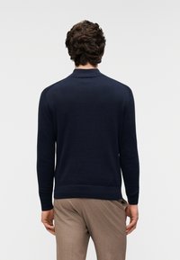 Pull en maille bleu marine avec col et poignets côtelés. Il présente un design élégant et une texture douce, porté avec un pantalon beige.